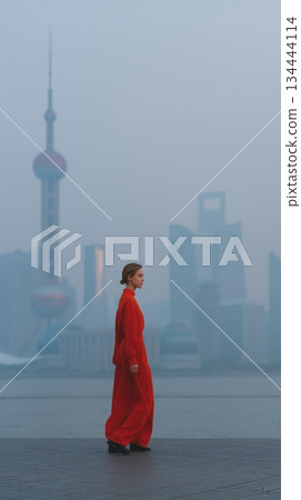 Woman in a vibrant red dress walks the pudong waterfront, gazing at the mist shrouded shanghai skyline with oriental pearl tower and futuristic skyscrapers looming through haze 134444114