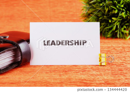 A card displaying Leadership sits against an orange backdrop with a book, a clip, and plant adding to the inspirational and motivational scene A card displaying Leadership sits against an orange backdrop with a book, a clip, and plant adding to the inspirational and motivational scene 134444420