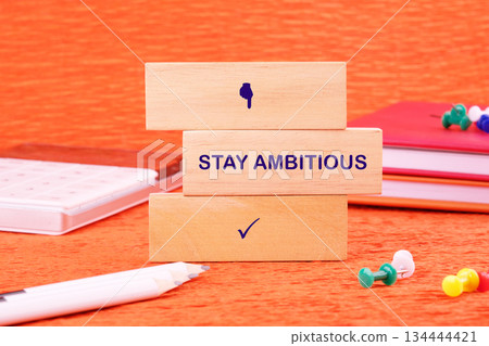 Stacked blocks inspire success with STAY AMBITIOUS. Notebooks, a pencil, and pushpins are neatly placed on a bright orange background suggesting a study or learning environment Stacked blocks inspire success with STAY AMBITIOUS. Notebooks, a pencil, and pushpins are neatly placed on a bright orange background suggesting a study or learning environment 134444421