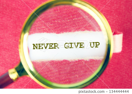 A message reading Never Give Up is magnified under a gold rimmed glass tool. The encouraging words sit on a small white piece of paper, set against a bright ruby red background 134444444