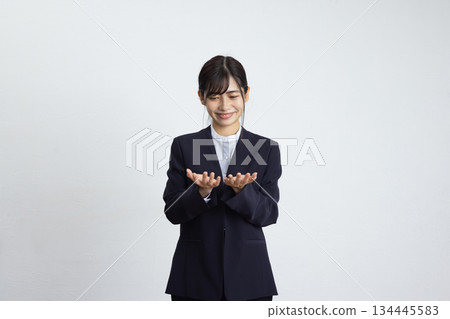 一位年輕的女商人面帶微笑，雙手放在胸前，站在一面白牆前。 134445583