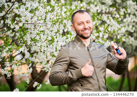 Man allergic using medical nasal drops, suffering from seasonal allergy at spring in blossoming garden. Handsome smiling man showing thumbs up near blooming tree outdoors. Spring allergy concept. Man allergic using medical nasal drops, suffering from seasonal allergy at spring in blossoming garden. Handsome smiling man showing thumbs up near blooming tree outdoors. Spring allergy concept. 134446270