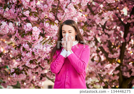 Woman allergic suffering from seasonal allergy at spring in blossoming garden at springtime. Woman sneezing and blowing nose using nasal handkerchief in front of blooming tree. Spring allergy concept. Woman allergic suffering from seasonal allergy at spring in blossoming garden at springtime. Woman sneezing and blowing nose using nasal handkerchief in front of blooming tree. Spring allergy concept. 134446275