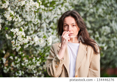 Woman allergic using medical nasal drops, suffering from seasonal allergy at spring in blossoming garden. Young woman treating runny nose in front of blooming tree outdoors. Spring allergy concept. Woman allergic using medical nasal drops, suffering from seasonal allergy at spring in blossoming garden. Young woman treating runny nose in front of blooming tree outdoors. Spring allergy concept. 134446276