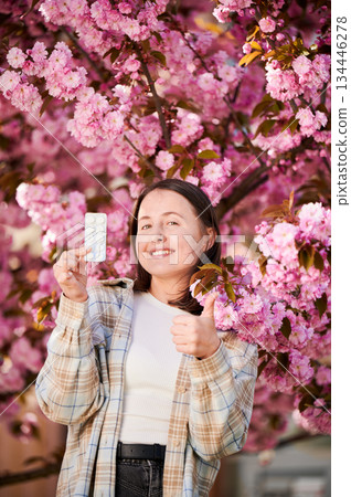 Woman allergic suffering from seasonal allergy at spring. Young happy woman showing allergy drugs, giving thumbs up, posing in blossoming garden at springtime. Antihistamine medication concept Woman allergic suffering from seasonal allergy at spring. Young happy woman showing allergy drugs, giving thumbs up, posing in blossoming garden at springtime. Antihistamine medication concept 134446278