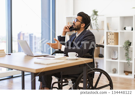 An Indian businessman in a wheelchair is talking on the phone while working in his office An Indian businessman in a wheelchair is talking on the phone while working in his office 134448122