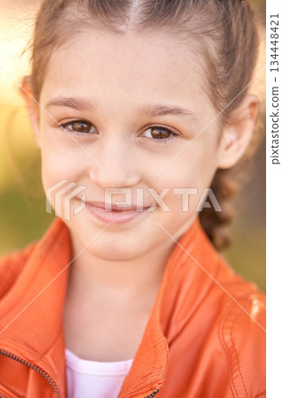 Smiling caucasian young girl in orange jacket outdoors with blurred background 134448421