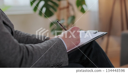 Close up man psychologist writing observation notes during therapy session with patient sitting beside in clinic room for mental health counseling support. Close up man psychologist writing observation notes during therapy session with patient sitting beside in clinic room for mental health counseling support. 134448596