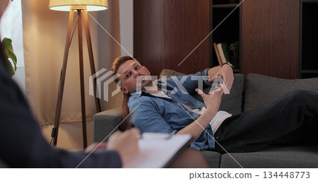 Man lies on couch sharing personal life problems in therapy room. Woman psychologists takes notes in notebook documenting key insights during conversation. Man lies on couch sharing personal life problems in therapy room. Woman psychologists takes notes in notebook documenting key insights during conversation. 134448773