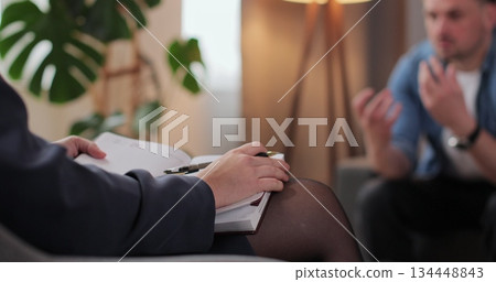 Close up woman psychologist writing observation notes during therapy session with man patient sitting beside in clinic room for mental health counseling support. 134448843