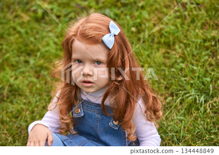 Caucasian young female child with red hair and blue bow sitting on green grass 134448849