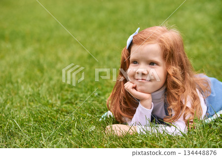 Red-haired caucasian young girl lying on grass with blue bow and thoughtful Red-haired caucasian young girl lying on grass with blue bow and thoughtful 134448876