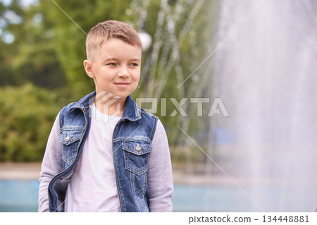 Caucasian young male child in denim jacket standing outdoors by a fountain Caucasian young male child in denim jacket standing outdoors by a fountain 134448881