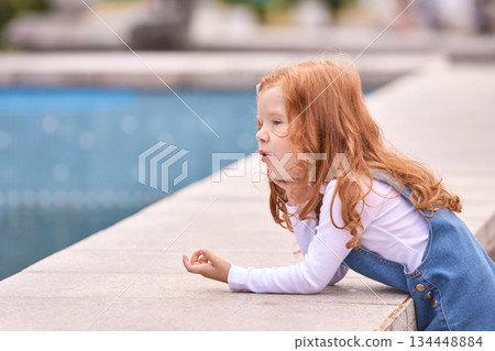 Young caucasian female child relaxing by poolside in denim dress with red hair Young caucasian female child relaxing by poolside in denim dress with red hair 134448884