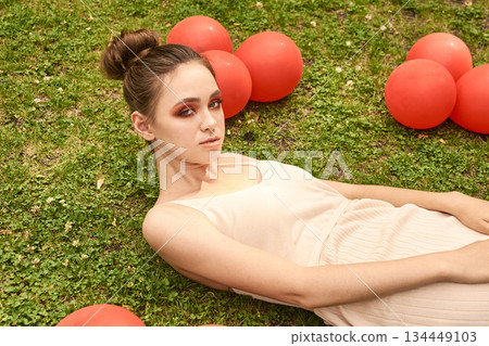 Young caucasian female relaxing on grass with red balloons in outdoor setting 134449103