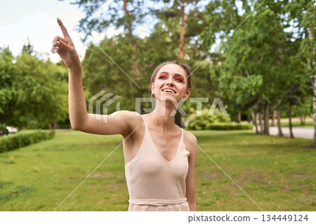 Caucasian young female smiling and pointing upwards in green park setting Caucasian young female smiling and pointing upwards in green park setting 134449124