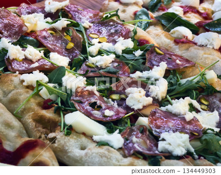 Fresh pizza topped with salami, cheese, and greens served on a wooden table in a busy restaurant Fresh pizza topped with salami, cheese, and greens served on a wooden table in a busy restaurant 134449303