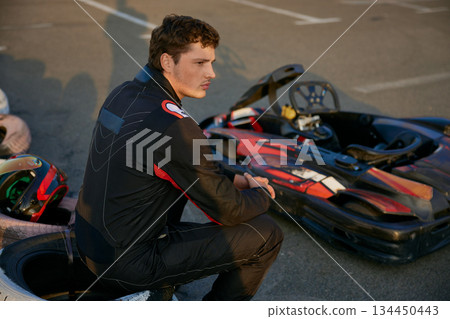 A focused young man in a racing suit sits beside his go-kart, contemplating the upcoming race A focused young man in a racing suit sits beside his go-kart, contemplating the upcoming race 134450443