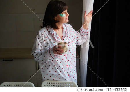 Woman wearing pajamas and eye patches enjoying warm drink while looking through window Woman wearing pajamas and eye patches enjoying warm drink while looking through window 134453752