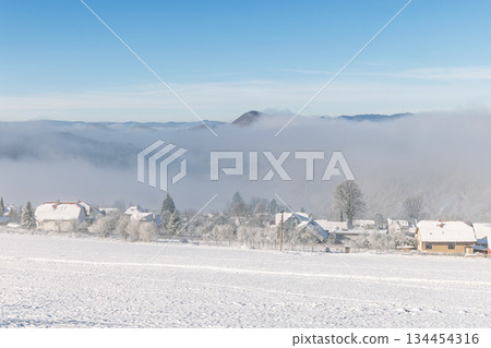Serene winter vista: A snow-covered village nestled beneath a blanket of fog, with hills visible in the distance under a clear blue sky. Peaceful winter scene. North of Slovakia, Europe. 134454316
