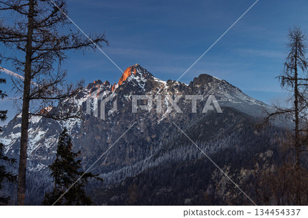 Lomnicky peak in High Tatras National Park, Slovakia, Europe. The sun's golden rays grace the mountain peak, creating a stunning display of light and shadow on the snow-dusted landscape. Lomnicky peak in High Tatras National Park, Slovakia, Europe. The sun's golden rays grace the mountain peak, creating a stunning display of light and shadow on the snow-dusted landscape. 134454337