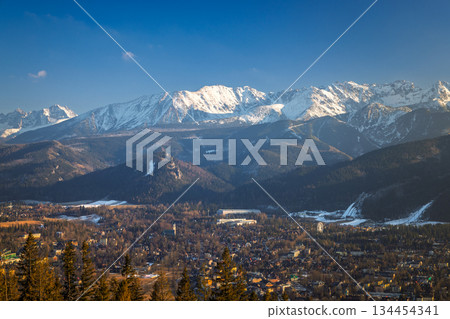 Picturesque view of a town nestled at the base of the mountains, capped with snow under a bright blue sky. Zakopane town at the foot of the Tatra Mountains in Poland, Europe. Picturesque view of a town nestled at the base of the mountains, capped with snow under a bright blue sky. Zakopane town at the foot of the Tatra Mountains in Poland, Europe. 134454341