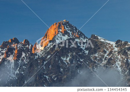 Lomnicky peak in High Tatras National Park, Slovakia, Europe. Golden hour bathes a majestic, snow-dusted mountain peak under clear blue skies, a serene and captivating alpine vista. 134454345