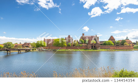 The Malbork Castle, a brick Gothic castle in northern Poland, Europe. Historical castle complex beside a reflective river under a bright sky with fluffy clouds on a sunny day. Medieval architecture. 134454354