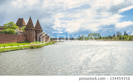 The Malbork Castle, a brick Gothic castle in northern Poland, Europe. Riverside brick castle with tall towers and trees under a cloudy sky casts reflections over the water on a sunny day. The Malbork Castle, a brick Gothic castle in northern Poland, Europe. Riverside brick castle with tall towers and trees under a cloudy sky casts reflections over the water on a sunny day. 134454358