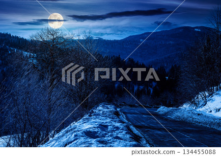 country road winding through winter landscape under dark sky at night. mountains in full moon light. travel background with pass and snow covered slopes. finance investment or abstract concepts 134455088