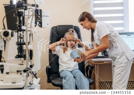 Holding mirror, looking at new eyewear. Little girl at the ophthalmologist clinic with doctor Holding mirror, looking at new eyewear. Little girl at the ophthalmologist clinic with doctor 134455358