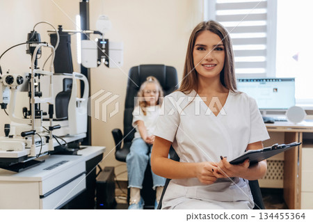 With notepad in hands. Little girl at the ophthalmologist clinic with doctor 134455364