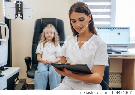 With notepad in hands. Little girl at the ophthalmologist clinic with doctor 134455367