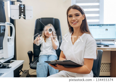 Sitting and holding notepad. Little girl at the ophthalmologist clinic with doctor Sitting and holding notepad. Little girl at the ophthalmologist clinic with doctor 134455374