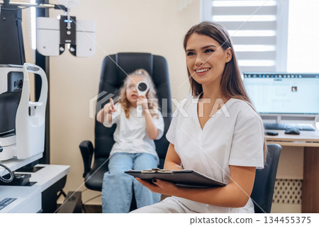 Sitting and holding notepad. Little girl at the ophthalmologist clinic with doctor Sitting and holding notepad. Little girl at the ophthalmologist clinic with doctor 134455375