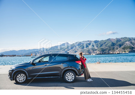 一位長髮年輕女子靠在冬日海邊的汽車旁，頭頂是蔚藍的天空。 134455458