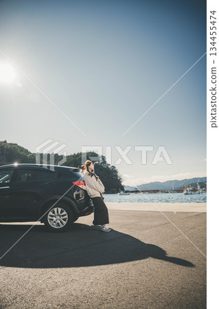 一位長髮年輕女子靠在冬日海邊的汽車旁，頭頂是蔚藍的天空。 134455474