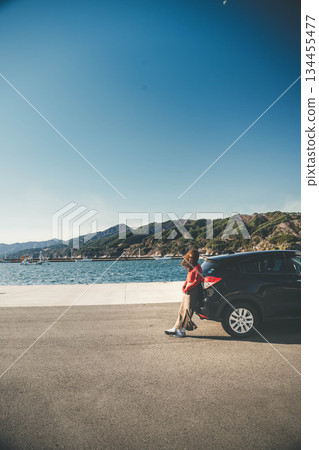 一位長髮年輕女子靠在冬日海邊的汽車旁，頭頂是蔚藍的天空。 134455477