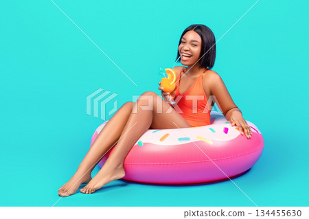 Young black lady wearing trendy swimsuit, sitting in inflatable ring, drinking fruit cocktail on blue background, copy space. Sexy woman with pool float and refreshing drink ready for beach party Young black lady wearing trendy swimsuit, sitting in inflatable ring, drinking fruit cocktail on blue background, copy space. Sexy woman with pool float and refreshing drink ready for beach party 134455630