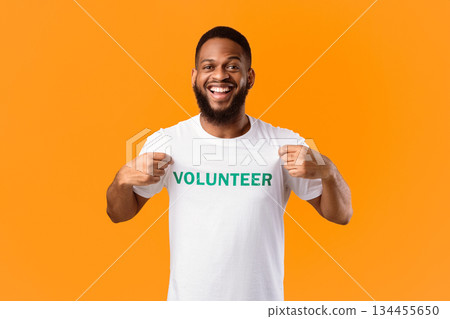 Volunteer's Portrait. Happy Black Activist Man Showing His T-Shirt With Volunteer Print Inscription On Chest Standing On Yellow Studio Background. Volunteering Job Concept 134455650