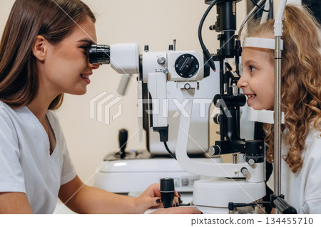 View from the side. Little girl at the ophthalmologist clinic with doctor View from the side. Little girl at the ophthalmologist clinic with doctor 134455710