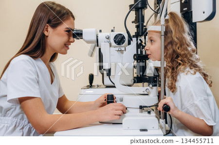 View from the side. Little girl at the ophthalmologist clinic with doctor View from the side. Little girl at the ophthalmologist clinic with doctor 134455711