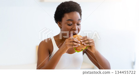 Cheat Meal Concept. Hungry Black Female Eating Big Burger Standing In Modern Kitchen At Home. Unhealthy Junk Food. Overeating And Weight Gain Problem, Eating Disorder And Nutrition 134455722