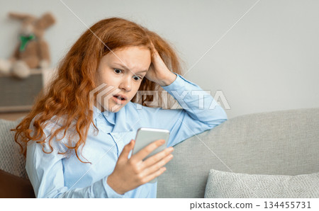 Shocked Surprised Young Teen Girl With Cellphone Reading Bad News Message Touching Head In Amazement Sitting On The Couch At Home. Confused Mobile Phone User, Call From Unknown Number 134455731