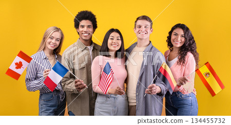 Student exchange, language learning, international holiday. Smiling multi ethnic student in casual, hold small flags of different countries, isolated on orange background, free space, studio shot 134455732
