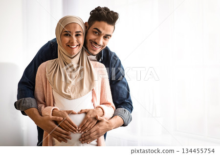 Portrait of happy pregnant religious muslim couple posing near window at home. Loving arab husband hugging his expecting wife in hijab from behind, making heart shaped sign on belly, copy space Portrait of happy pregnant religious muslim couple posing near window at home. Loving arab husband hugging his expecting wife in hijab from behind, making heart shaped sign on belly, copy space 134455754