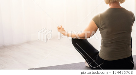 Back view of peaceful senior woman meditating in lotus pose at home, empty space. Calm mature lady doing morning yoga practice, facing window, making gyan mudra, reaching zen Back view of peaceful senior woman meditating in lotus pose at home, empty space. Calm mature lady doing morning yoga practice, facing window, making gyan mudra, reaching zen 134455768