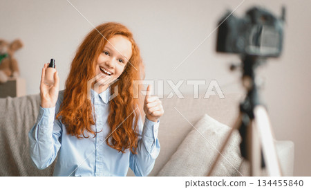 Beauty Advice And Blog. Smiling female teen doing makeup, recording cosmetics product review, showing her foundation for young skin to camera on tripod. Girl filming video, showing thumbs up 134455840