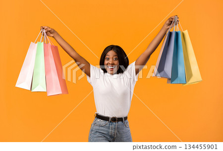 Emotional african american young woman raising hands up with colorful shopping bags and screaming cheerfully, celebrating sales season beginning over orange studio background 134455901