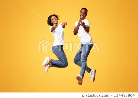 Happy black lovers attractive young man and woman wearing jeans and white t-shirts posing on orange studio background, jumping in the air and pointing fingers at camera. People lifestyle concept 134455904